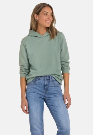 Kvinde iført en lysegrøn hættetrøje og blå jeans, smilende og kigger til siden med hænderne afslappet langs siderne.