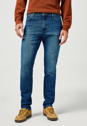 Blue denim jeans with a slim fit, featuring subtle fading and classic five-pocket design. Paired with tan sneakers and a brown sweatshirt.