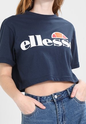 Camiseta corta de color navy hecha de algodón, con mangas cortas enrolladas y un atrevido texto de logo en blanco. Combinada con jeans de mezclilla de cintura alta.