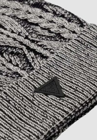 Berretto lavorato a maglia in nero e grigio, caratterizzato da un motivo a trecce testurizzato e da un risvolto a coste. È applicata una toppa triangolare con logo nero.