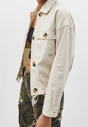 Beige blousejack met grote schildpadknopen gedragen over een zwart cropped topje, gecombineerd met groene en bruine camouflage cargobroeken.