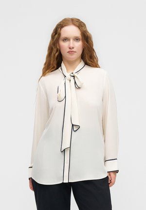 Γυναίκα με μακριά σγουρά μαλλιά φοράει μια κρεμ μπλούζα με μαύρη περίμετρο και δεμένη γραβάτα στο λαιμό, σε συνδυασμό με μαύρο παντελόνι, στέκεται μπροστά σε λευκό φόντο.