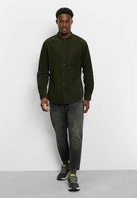 Dunkelgrünes Hemd mit Stehkragen, einer Brusttasche und knöpfbaren Manschetten, kombiniert mit grauen Jeans und grünen Sneakers.