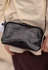 Borsa a tracolla nera in pelle con texture coccodrillo, forma rettangolare e chiusura a zip. Presenta una tracolla rimovibile e dettagli in metallo dorato.