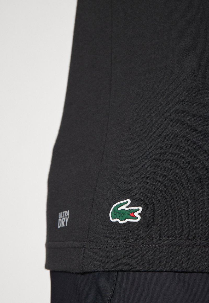 Camisa negra de textura suave, con un logo de cocodrilo en verde y blanco y el texto "ULTRA DRY" en la parte inferior izquierda.