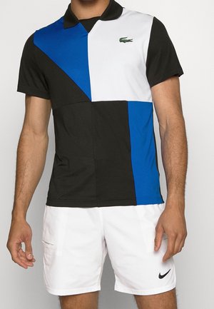 Mężczyzna w czarno-niebiesko-białej koszulce polo z blokowym wzorem i logo Lacoste oraz białych szortach Nike z czarnym logo Swoosh.