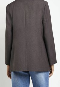 Hnědý blazer se strukturovaným designem, s dlouhými rukávy, rovnoměrným střihem a hladkou texturou. Nesený s světle modrými džíny.