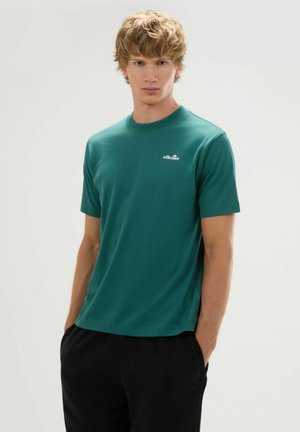 Junger Mann mit blondem Haar, der ein grünes ellesse T-Shirt und schwarze Hose trägt, steht mit den Händen in den Taschen vor einem schlichten Hintergrund.
