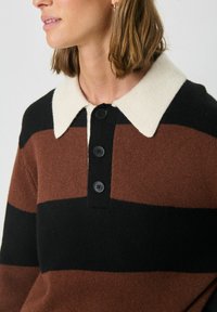 Strikket pullover med sorte, brune og creme horisontale striber. Har en cremefarvet krave og tre sorte knapper. Blød tekstur.