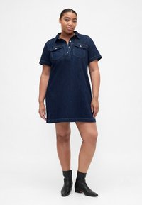 Temno modra denim obleka s kratkimi rokavi, dvema sprednjima žepoma in gumbe na sprednji strani, kombinirana s črnimi gležnjarji.