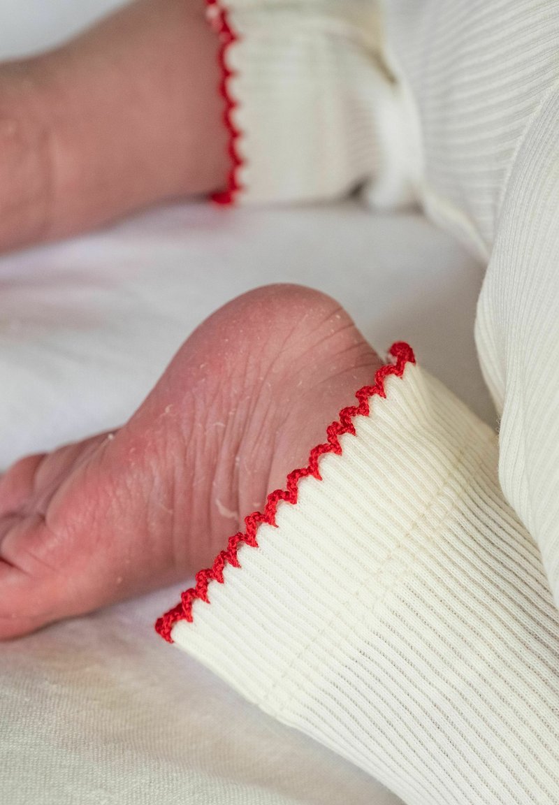 Gros plan sur les pieds nus d’un bébé portant un pantalon blanc côtelé avec des bords festonnés rouges, reposant sur une surface en tissu clair.