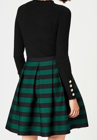 Zwarte geribbelde langengemouwen top gecombineerd met een groen en zwart gestreepte skaterrok, voorzien van een aansluitende taille en gouden knopaccenten op de mouwen.