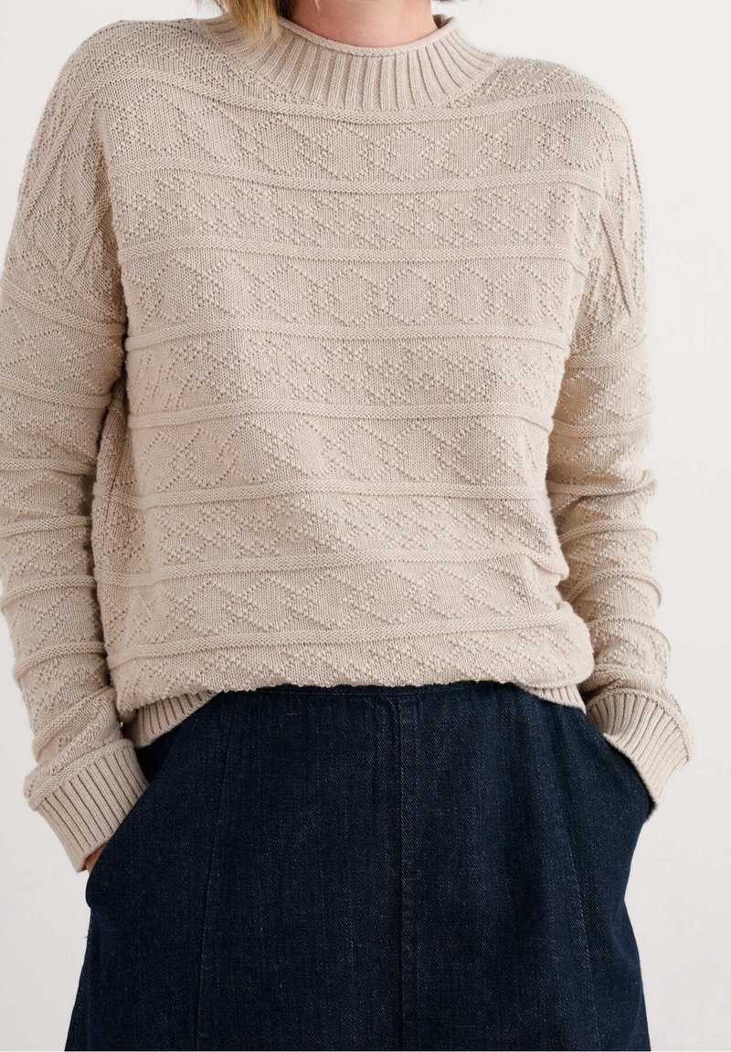 Pull en maille beige texturée avec un motif en losange, col rond et poignets côtelés, porté sur un pantalon bleu foncé avec les mains dans les poches.