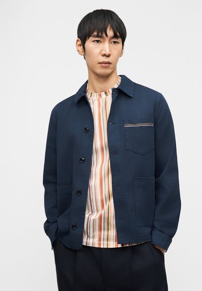 Νέος άνδρας που φοράει ένα σκούρο μπλε σακάκι με κουμπιά πάνω από ένα πολύχρωμο πουκάμισο με κάθετες ρίγες, στέκεται με τα χέρια στις τσέπες μπροστά από απλό φόντο.