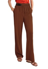 Bruna vida byxor med texturerad yta, med framfickor och ett svart bälte. Tillsammans med bruna sandaler, vilket ger en avslappnad look.