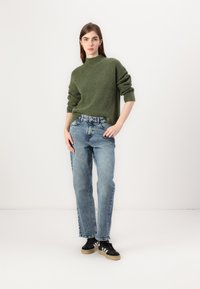Groene gebreide trui met een hoge hals, gecombineerd met vervaagde blauwe rechte jeans en zwarte schoenen met beige zolen. Eenvoudig ontwerp en textuur.