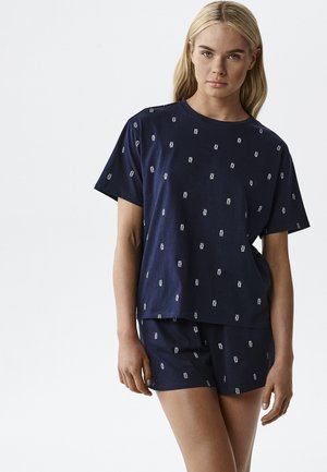 Młoda kobieta o długich blond włosach, ubrana w dopasowaną granatową koszulkę z krótkim rękawem i spodenki z małym białym wzorem tekstowym.