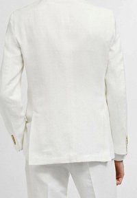 Uomo che indossa un abito bianco su misura con bottoni dorati sulle maniche, visto da dietro su uno sfondo chiaro e semplice.