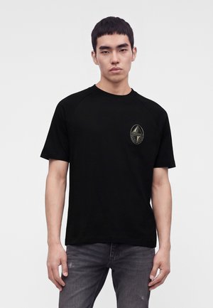 Homme portant un T-shirt noir à manches courtes avec un petit logo Hugo sur la poitrine gauche et un jean gris foncé sur un fond uni.