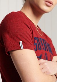 Camiseta roja de manga corta con superficie texturizada, estampado gráfico azul y etiqueta de logo en el dobladillo. Diseño simple de cuello redondo.