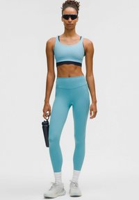 Gaiši zils sporta krūšturis un augsto jostasvietu legingi gludā audumā. Modelis tur melnu ūdens pudeli, valkājot tumšas saulesbrilles un gaišas kedas.