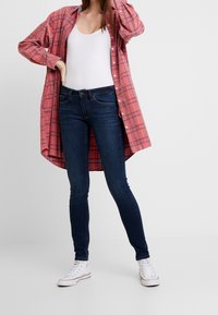 Röd rutmönstrad skjorta med knapp över en vit linne, kombinerad med mörkblå tajta jeans och vita högskaftade sneakers.