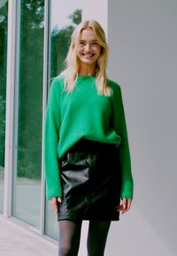 Žalias pūkuotas megztinis su ilgomis rankovėmis, derinamas su juodu odiniu mini sijonu. Lygus paviršius ir ryški spalvų kontrastas aprangoje.