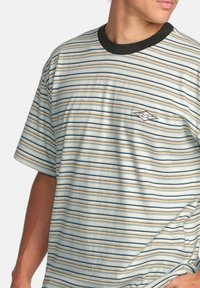 Randig T-shirt i ljusblått, kräm och mörkblått, med en svart rundad halsringning och en broderad logotyp på bröstet.