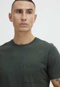 Camiseta de manga corta verde oscuro hecha de tela ligera, con un bolsillo en el pecho y un clásico cuello redondo. Textura suave.