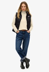 Schwarzweste mit Polsterung über einem cremefarbigen Rollkragenpullover, kombiniert mit lockeren blauen Jeans und schwarzen Schnürstiefeln, die einen strukturierten Gürtel und hochgekrempelte Hosenbeine aufweisen.