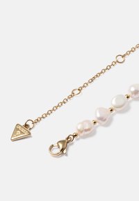 Pulseira dourada com pérolas brancas irregulares e fecho tipo lagosta. Acentuada com uma etiqueta triangular gravada com "GUESS."