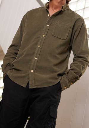 Homme portant une chemise en velours côtelé vert olive boutonnée et un pantalon cargo noir, les mains dans les poches, debout à l'intérieur près d'un mur blanc.