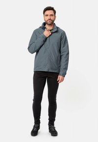 Giacca grigia impermeabile con cappuccio, cerniera frontale e polsini elasticizzati, abbinata a pantaloni neri e stivali neri. Design semplice e funzionale.