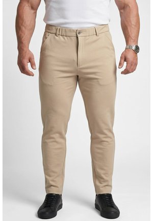 Hombre con pantalones ajustados beige, camiseta blanca sin mangas, zapatillas negras y reloj de pulsera plateado en la muñeca izquierda.