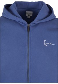Marineblå zip-up hoodie lavet af blødt stof. Har hætte med snøre og hvidt broderet logo på venstre bryst. Lynlås lukning.
