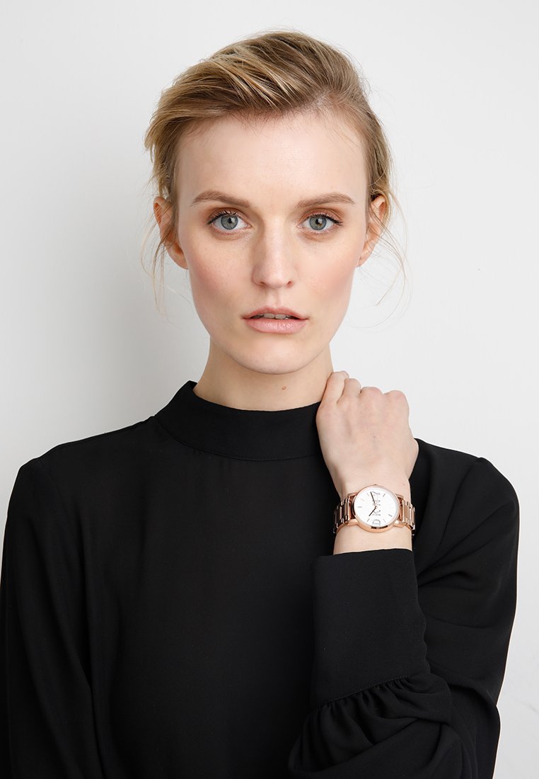 Orologio in metallo rosa con quadrante bianco, indicatori dell'ora sottili e cassa rotonda, indossato su un polso. Modella in una blusa nera a collo alto.