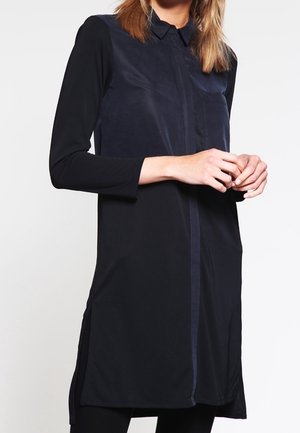 Woman wearing a long-sleeve black tunic with a shiny navy front panel and a buttoned collar, standing with hands lightly clasped.