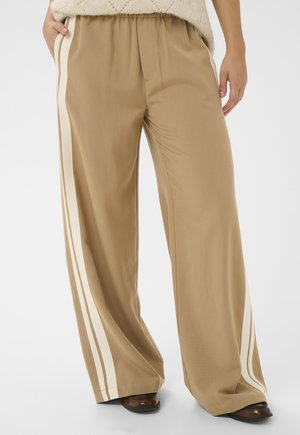 Beige bukser med vide ben og cremefarvede sidestriber, båret med brune sko og en tekstureret beige trøje, hænder afslappet ned langs siderne.