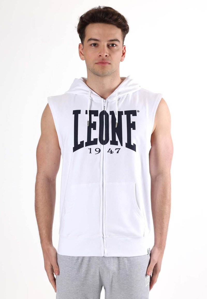 Jeune homme portant un sweat à capuche sans manches blanc zippé avec le texte "LEONE 1947" et un pantalon de survêtement gris clair, debout devant un fond uni.