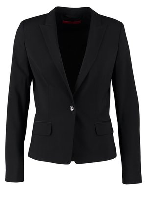 Femme portant un blazer noir, un haut en satin rose, un pantalon noir, des talons noirs, et tenant un sac à main noir.