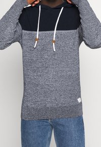 Stickad grå huvtröja med en marin panel över bröstet, vita snören, bruna accenter och strukturerad tyg, tillsammans med blå jeans.