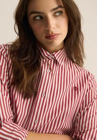 Koszula z guzikami, w czerwono-białe pionowe pasy, z kołnierzykiem, długimi rękawami i małym haftowanym logo na klatce piersiowej.