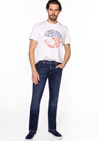 Biała koszulka z czerwono-niebieskim nadrukiem, zestawiona z ciemnoniebieskimi dżinsami. Model stoi swobodnie z rękami w kieszeniach, nosząc niebieskie trampki.