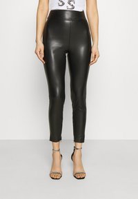 Leggings en simili cuir noir avec taille haute, coupe slim, zips aux chevilles et texture lisse et brillante. Portés avec des talons noirs à lanières.