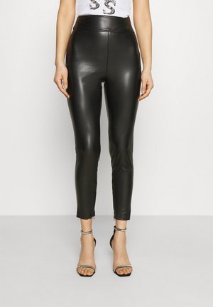 Leggings en simili cuir noir avec taille haute, coupe slim, zips aux chevilles et texture lisse et brillante. Portés avec des talons noirs à lanières.