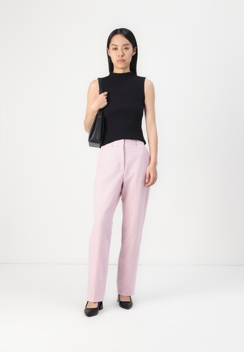 Schwarzes geripptes ärmelloses Oberteil, hellrosa taillierte Hose und schwarze hochhackige Schuhe. Das Model trägt eine kleine schwarze Handtasche.