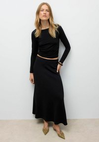 Donna con lunghi capelli biondi che indossa un top nero a maniche lunghe, una gonna maxi nera, un bracciale marrone e delle scarpe verdi oliva a punta, in piedi contro un muro bianco.