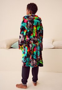 Fleece-Bademantel mit mehrfarbigem abstraktem Muster, einschließlich Fußballbällen, mit Kapuze und gebundenem Taillengürtel. Trägt schwarze Hosen mit weißen Streifen darunter.