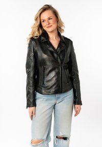 Schwarze Leder-Motorradjacke mit Kragen, Reißverschluss vorne, Stern-Patches auf den Ärmeln und strukturierten Akzenten. Kombiniert mit hellen, gewaschenen Jeans.