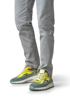 Baskets vertes et jaunes en mesh et suède, avec un motif camouflage sur la languette et une semelle texturée grise. Portées avec un jean gris.
